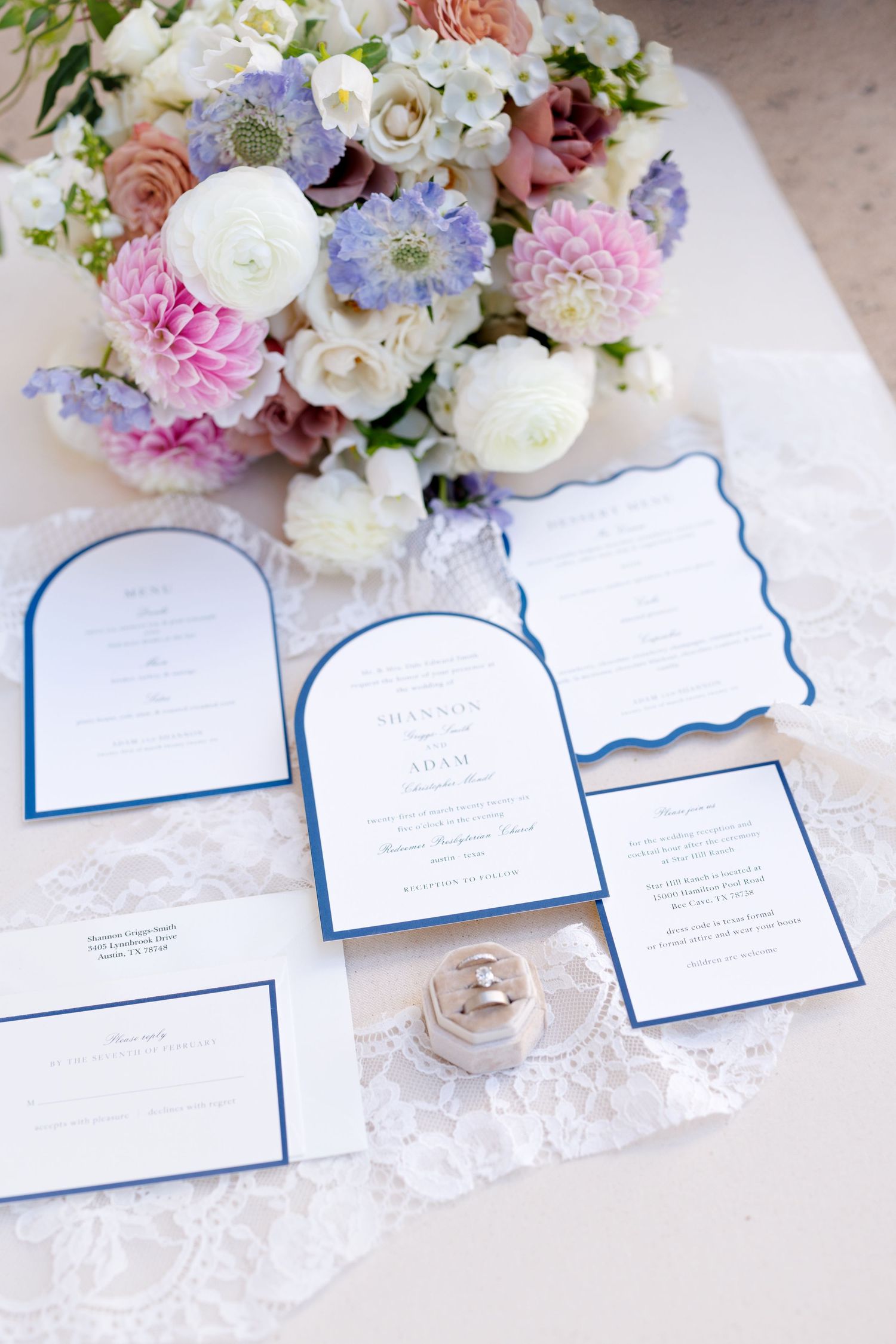 Elegant wedding invitation suite with blue borders displayed alongside pink and purple floral bouquet on lace tablecloth.