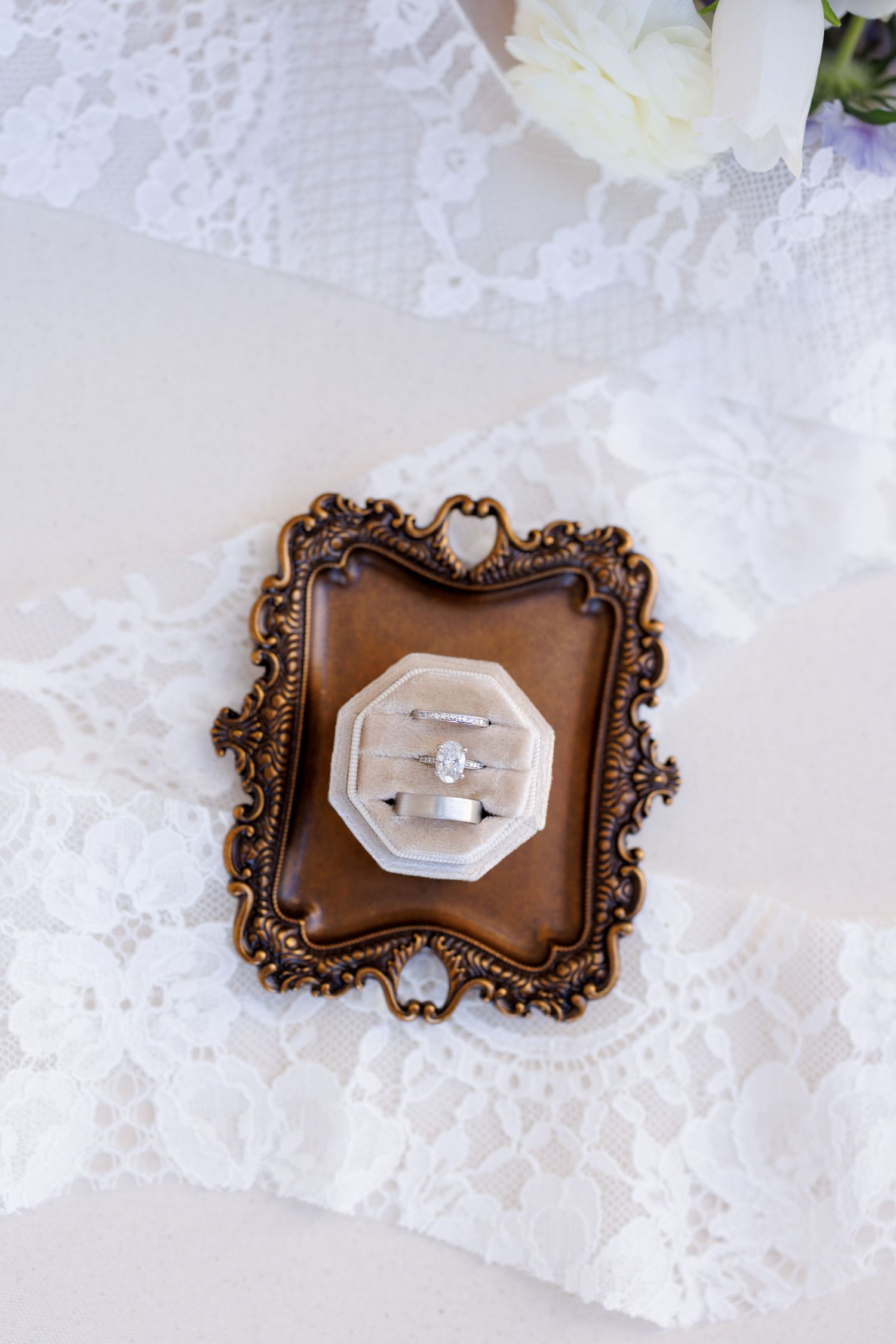 Ornate bronze-colored ring box with decorative frame design on white lace background.