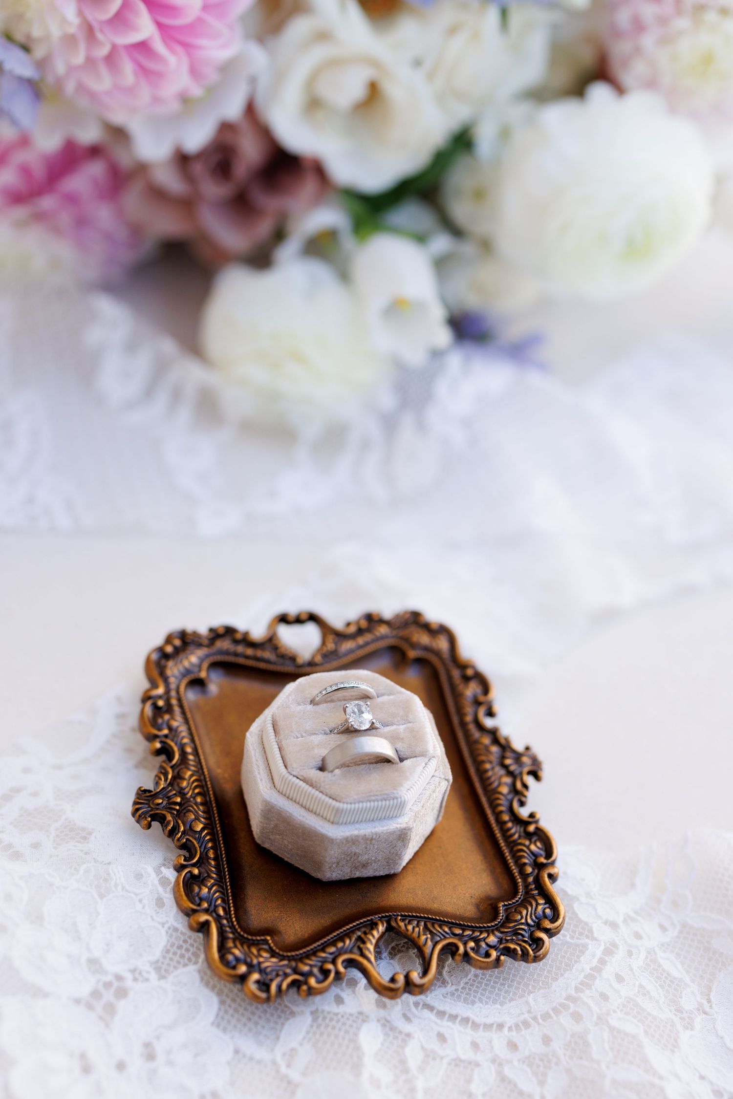 Elegant wedding ring display on bronze vintage-style holder with pink and white flowers in background.