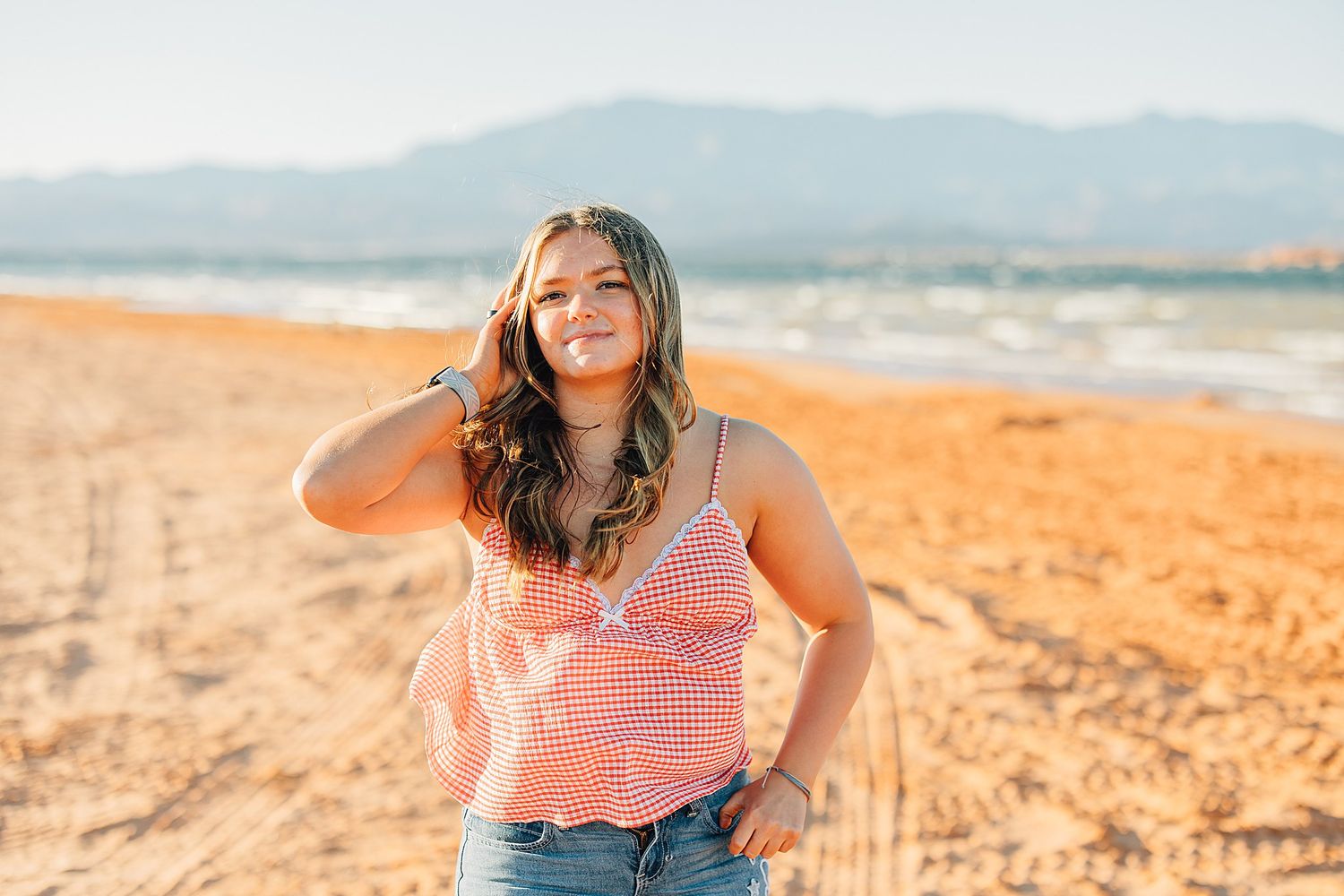 Young woman in pink top and jeans smiling on sandy beach with mountains and ocean in background.
