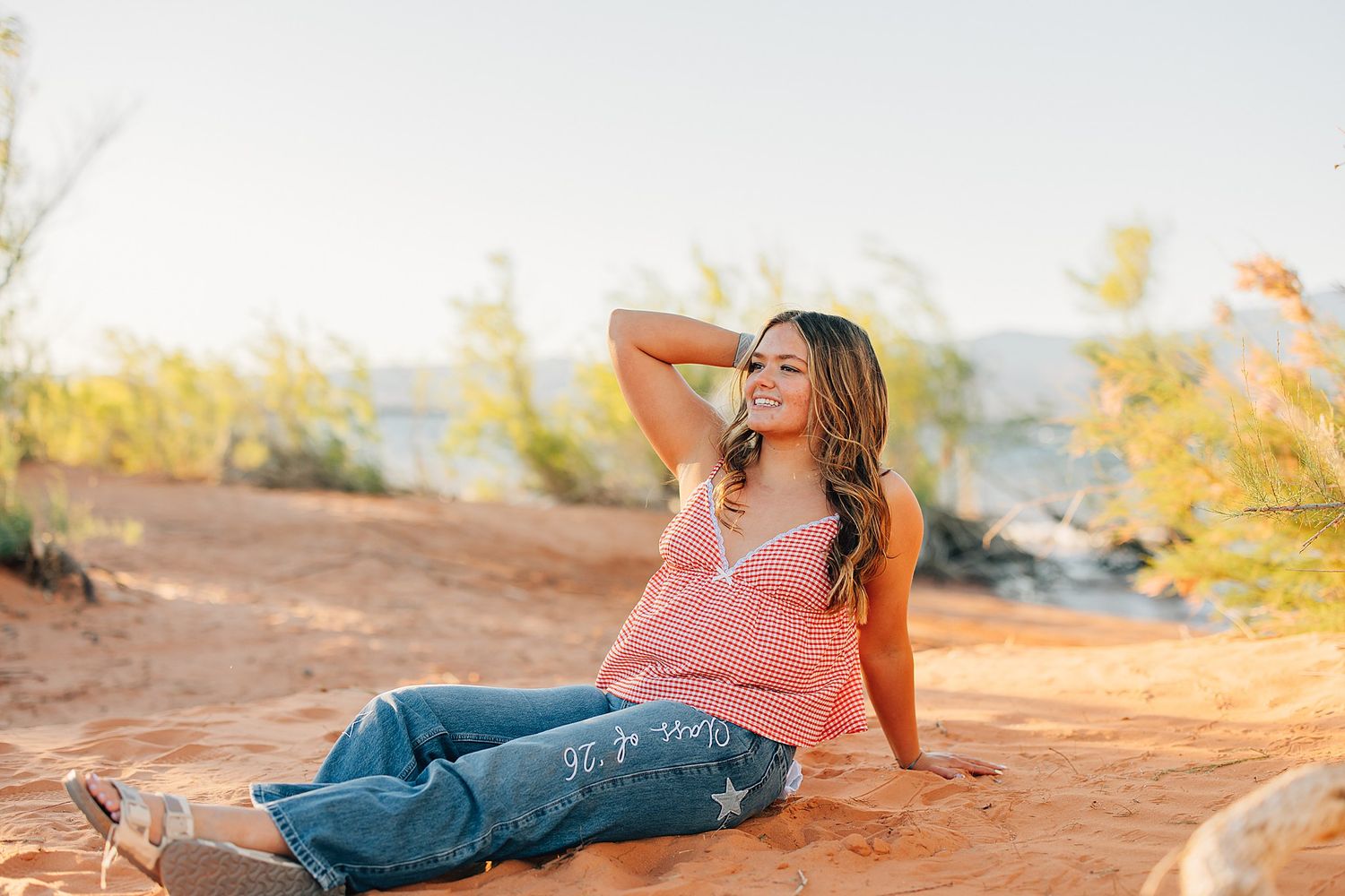 Young woman in pink top and jeans sitting on red sand, smiling outdoors in golden hour sunlight.