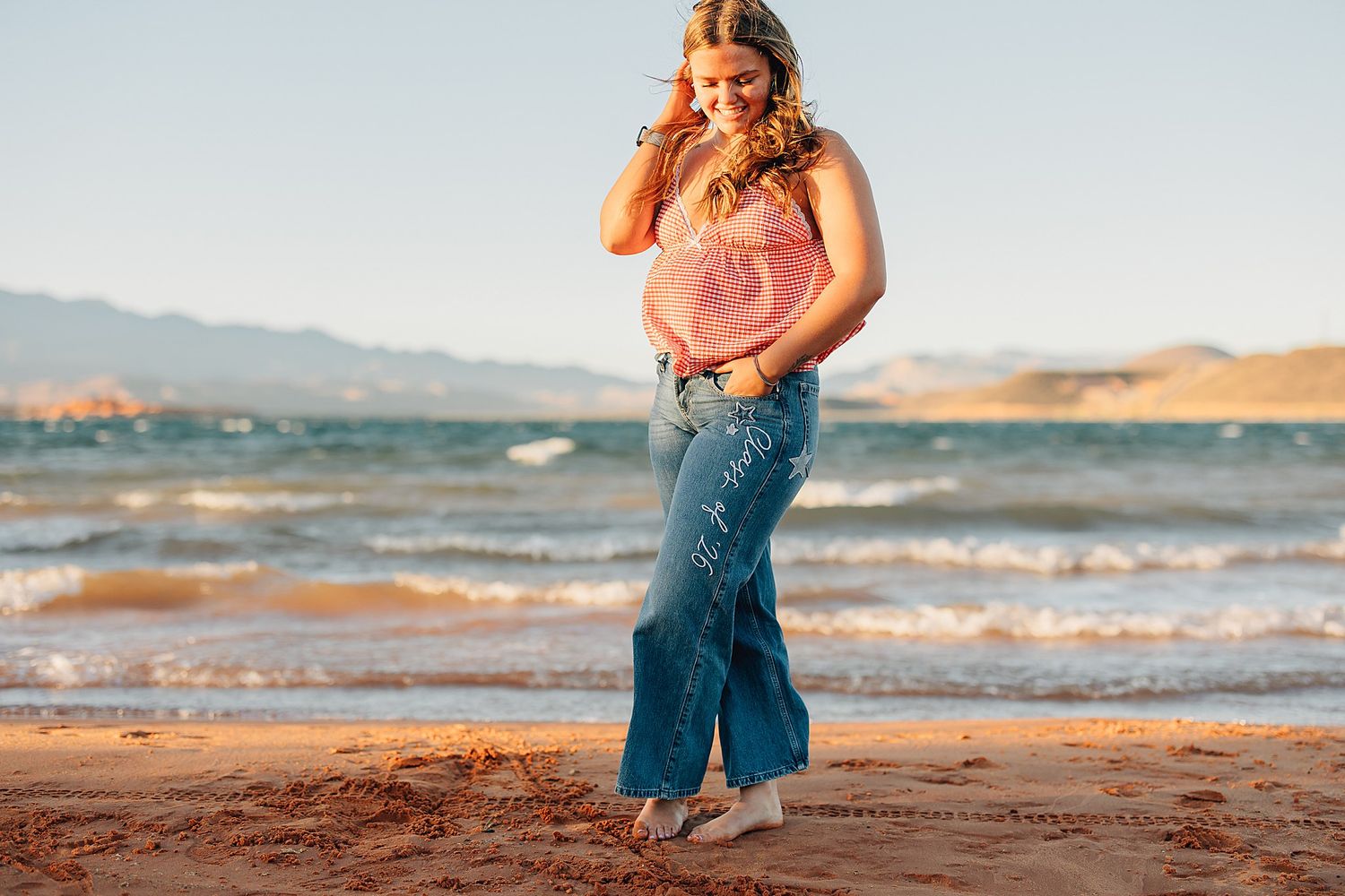 A woman in a pink top and wide-leg jeans poses on a sandy beach with ocean waves and mountains behind her.