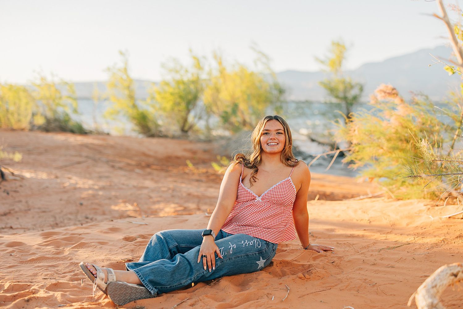 Young woman smiling while sitting on red sand near a lake with desert trees and mountains in the background.