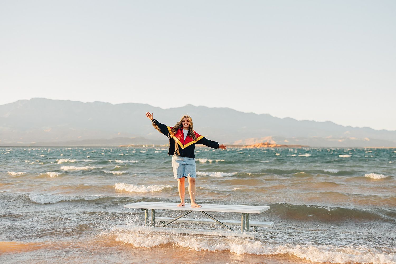 Graduate in cap and gown balancing on dock at beach with mountains in background.
