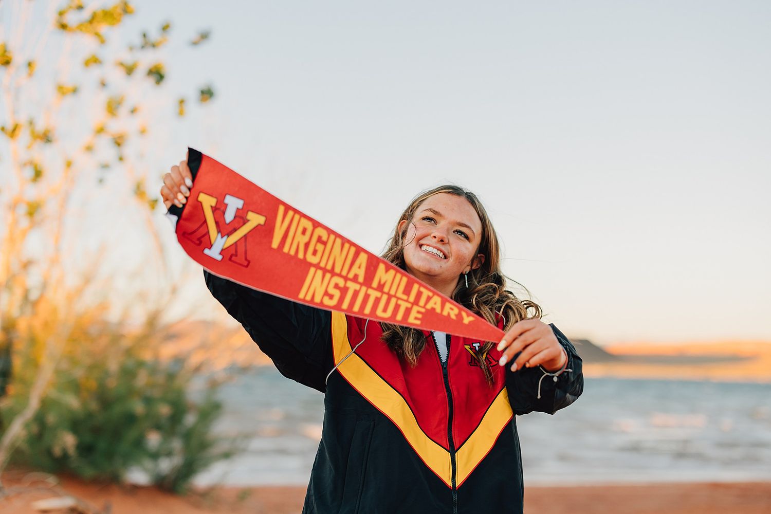 Graduate proudly holds Virginia Military Institute pennant near waterfront at sunset.