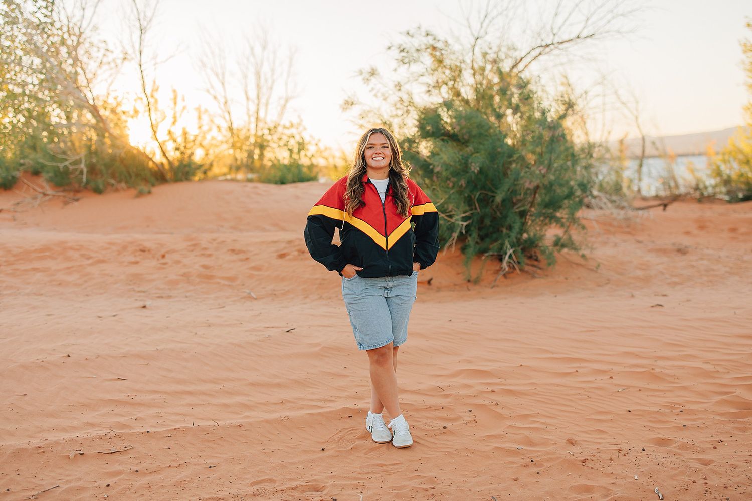 Young woman in red varsity jacket smiling on red sand dunes at golden hour.