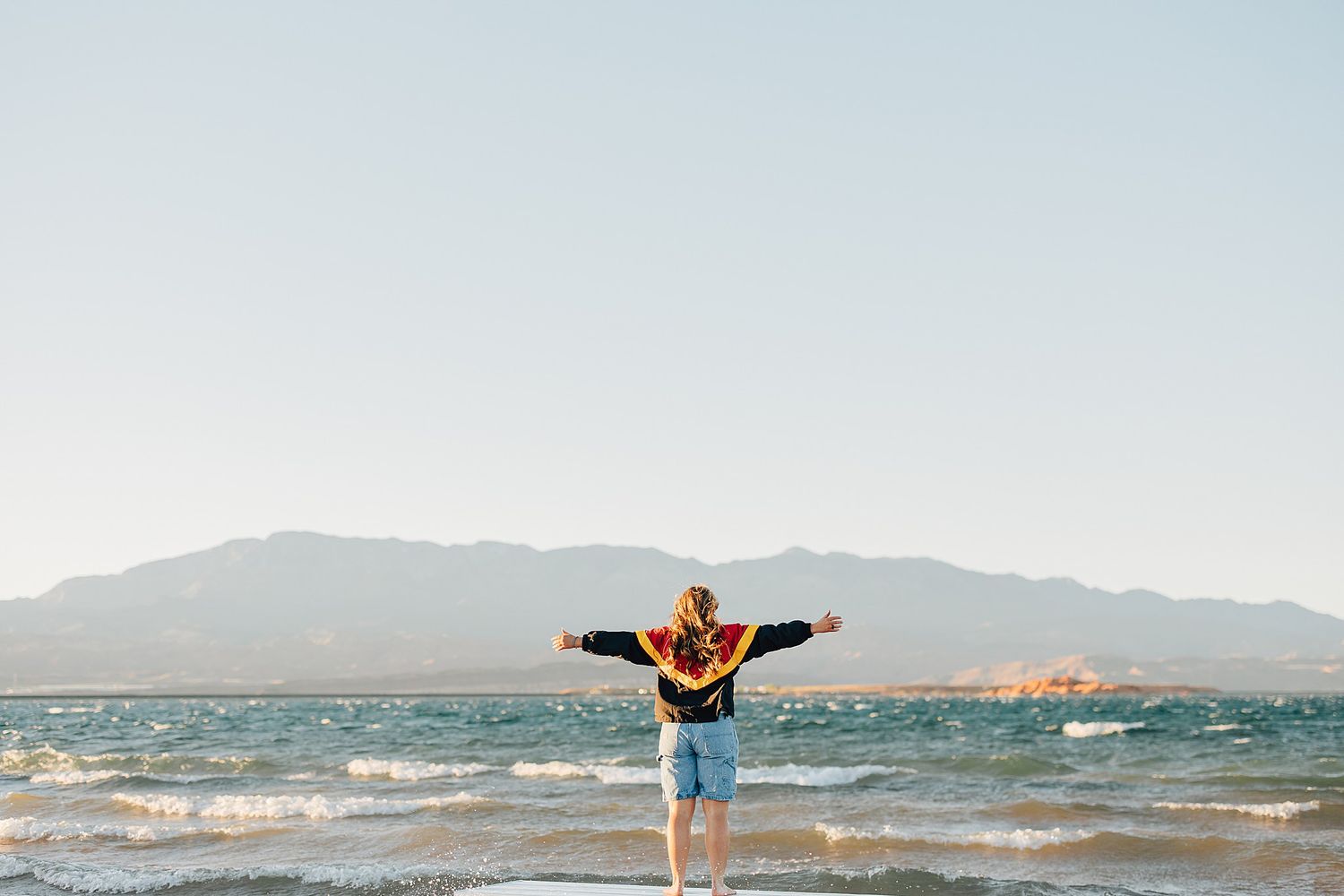 Person standing on a sunny beach with arms outstretched, facing the ocean with mountains in the background.