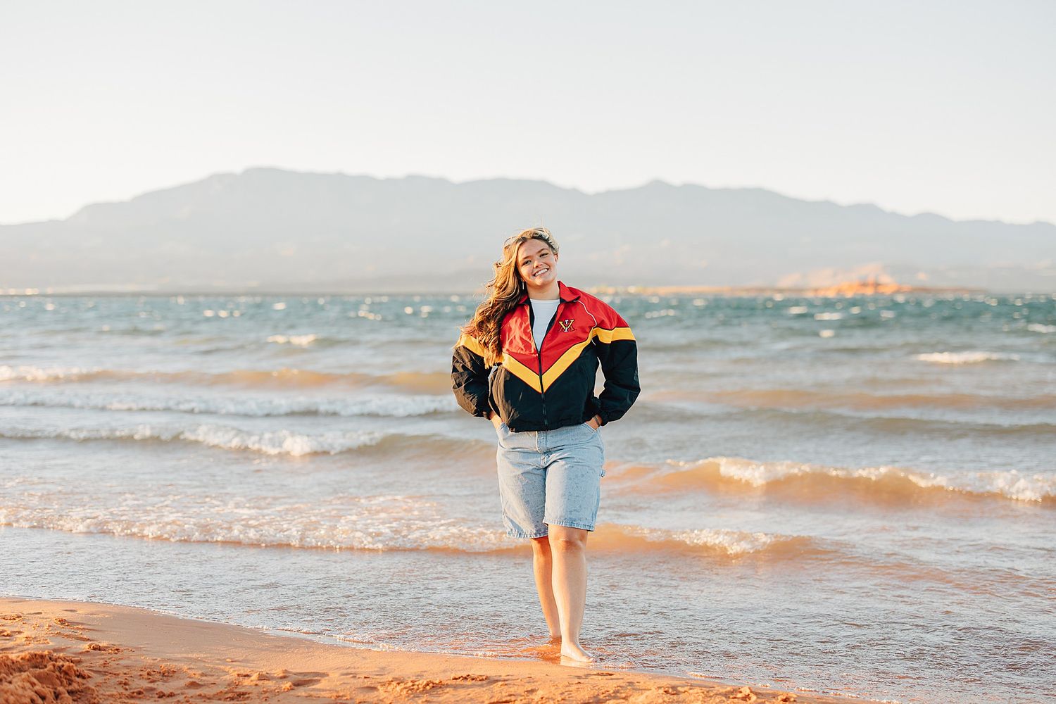 Woman in colorful windbreaker and denim shorts walking barefoot on sandy beach with mountains in background.