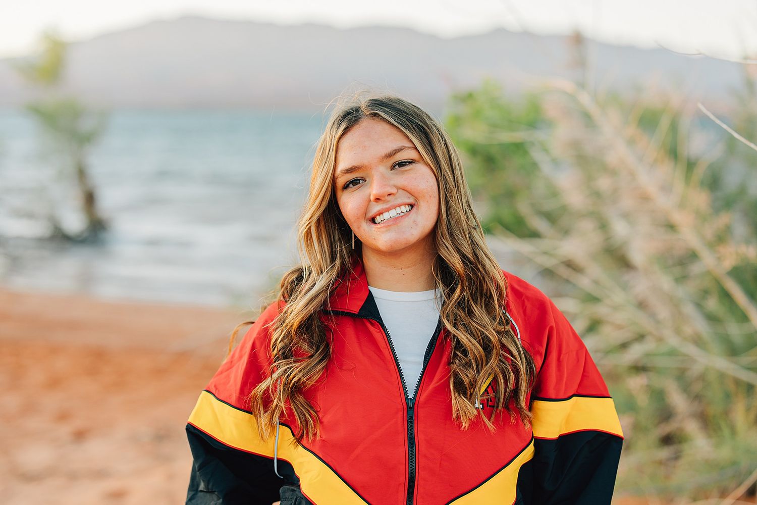 Smiling young woman with wavy hair wearing a red, yellow, and black jacket near a beach.