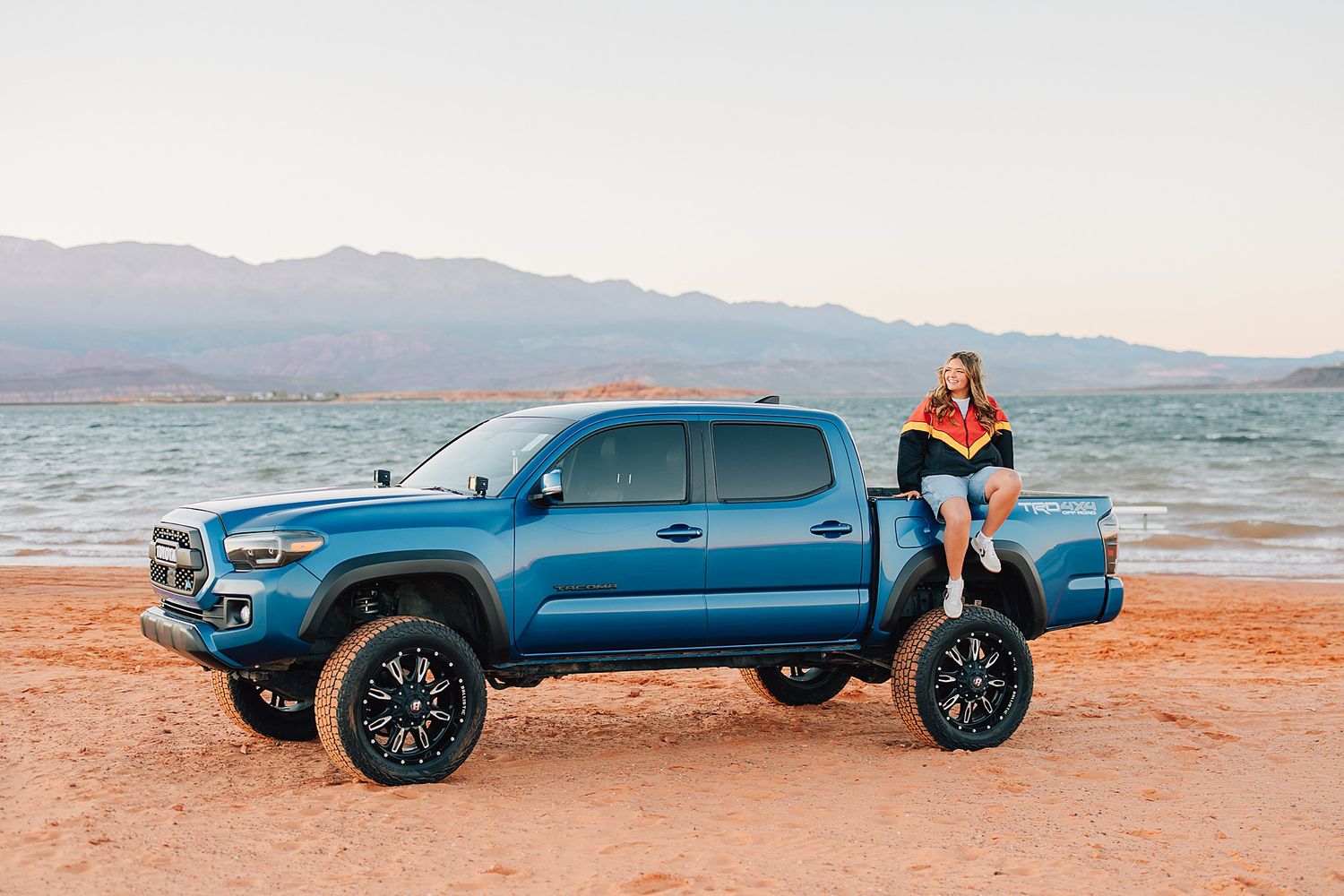 Woman sitting on blue Toyota Tacoma TRD truck bed at sandy desert lake shore with mountains in background.
