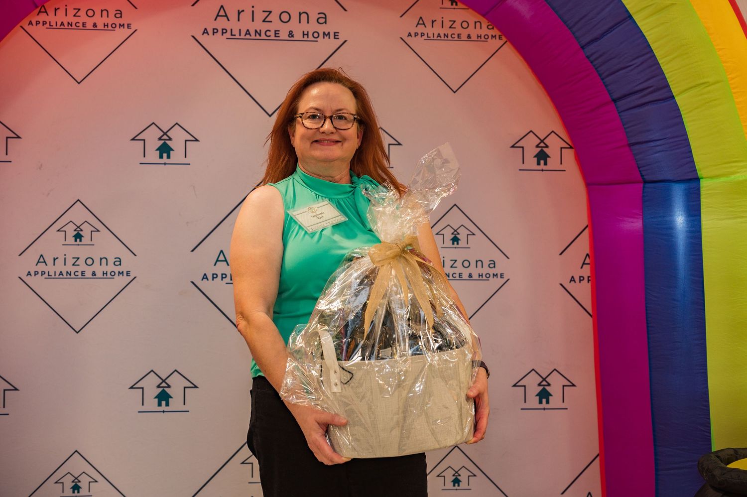 Woman in teal top holding a gift basket in front of Arizona Appliance and Home branded backdrop.