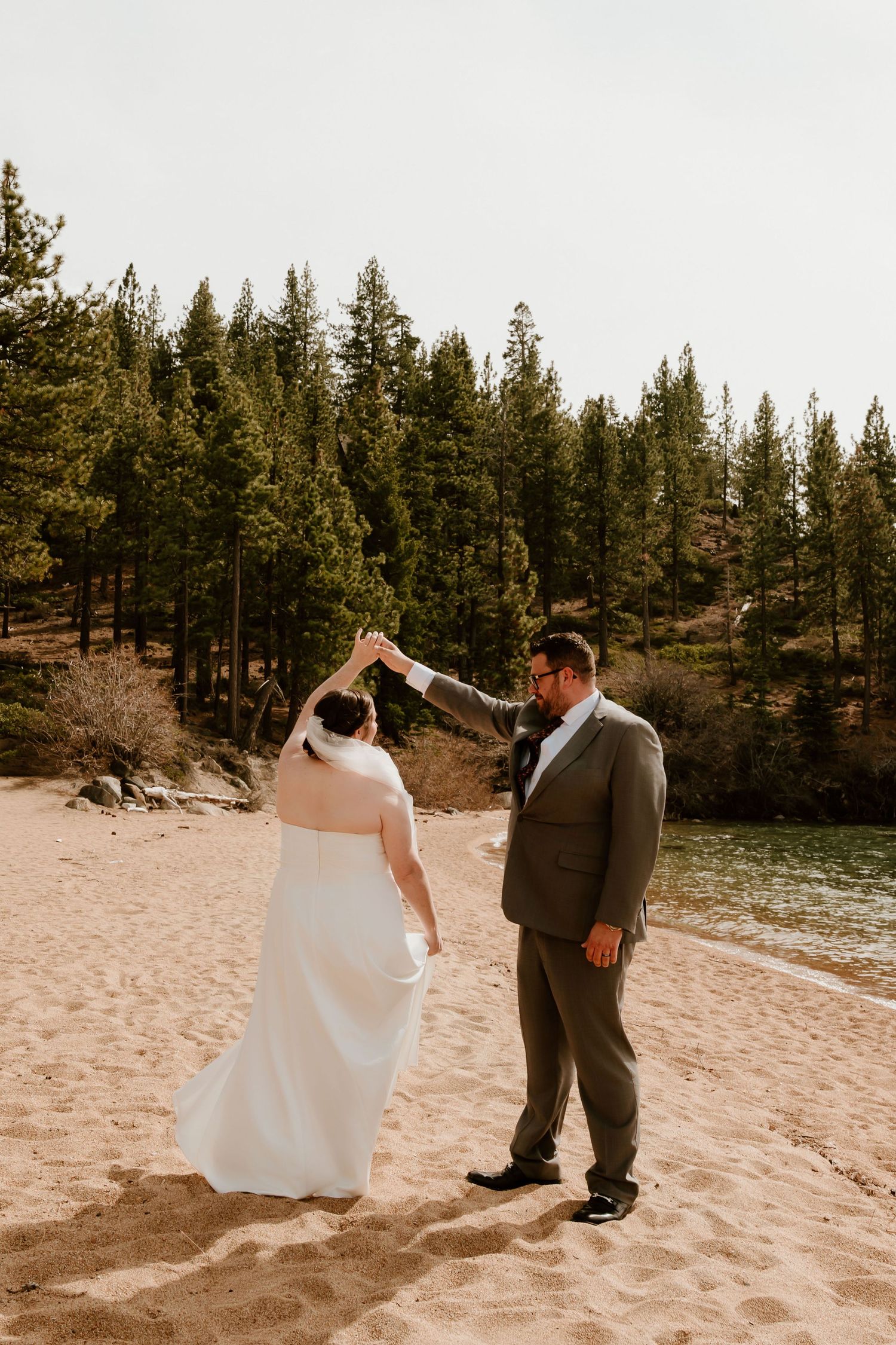 A couple shares a romantic moment on a sandy beach with evergreen trees in the background at Lake Tahoe.