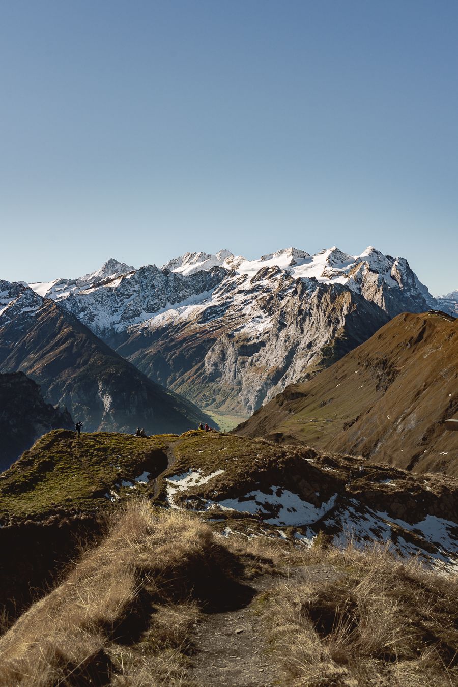 Hikers on a mountain trail with snow-dusted peaks and green valleys in the Swiss Alps.