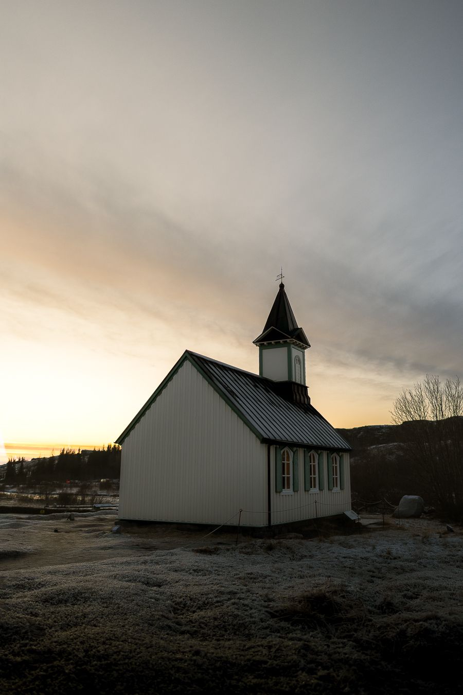 Small white wooden church with steeple silhouetted against colorful winter sunset sky in Iceland.