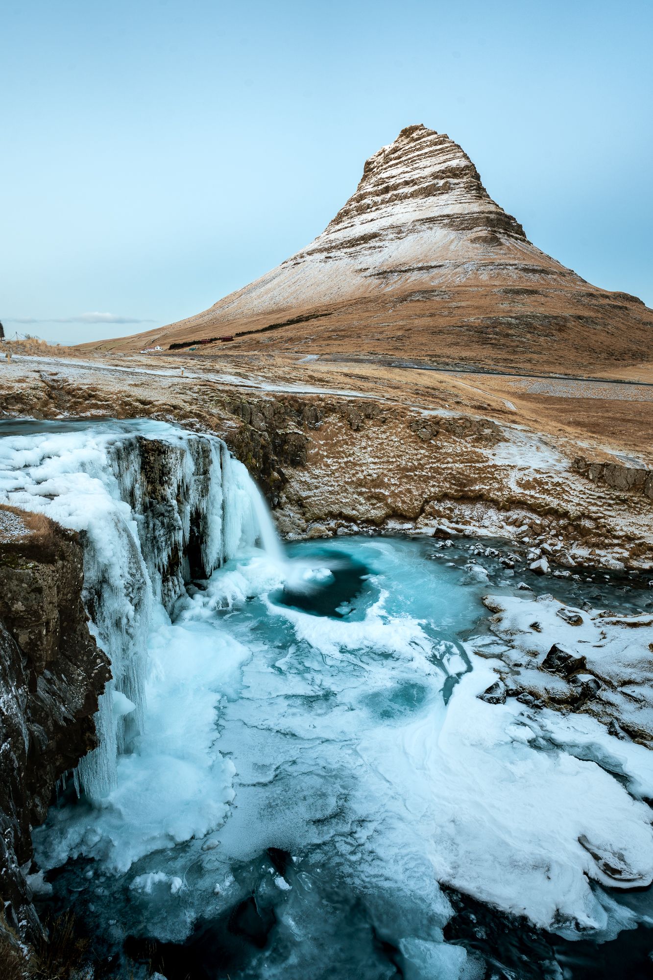 Iconic Kirkjufell mountain with frozen waterfall and icy blue river in Iceland's winter landscape.