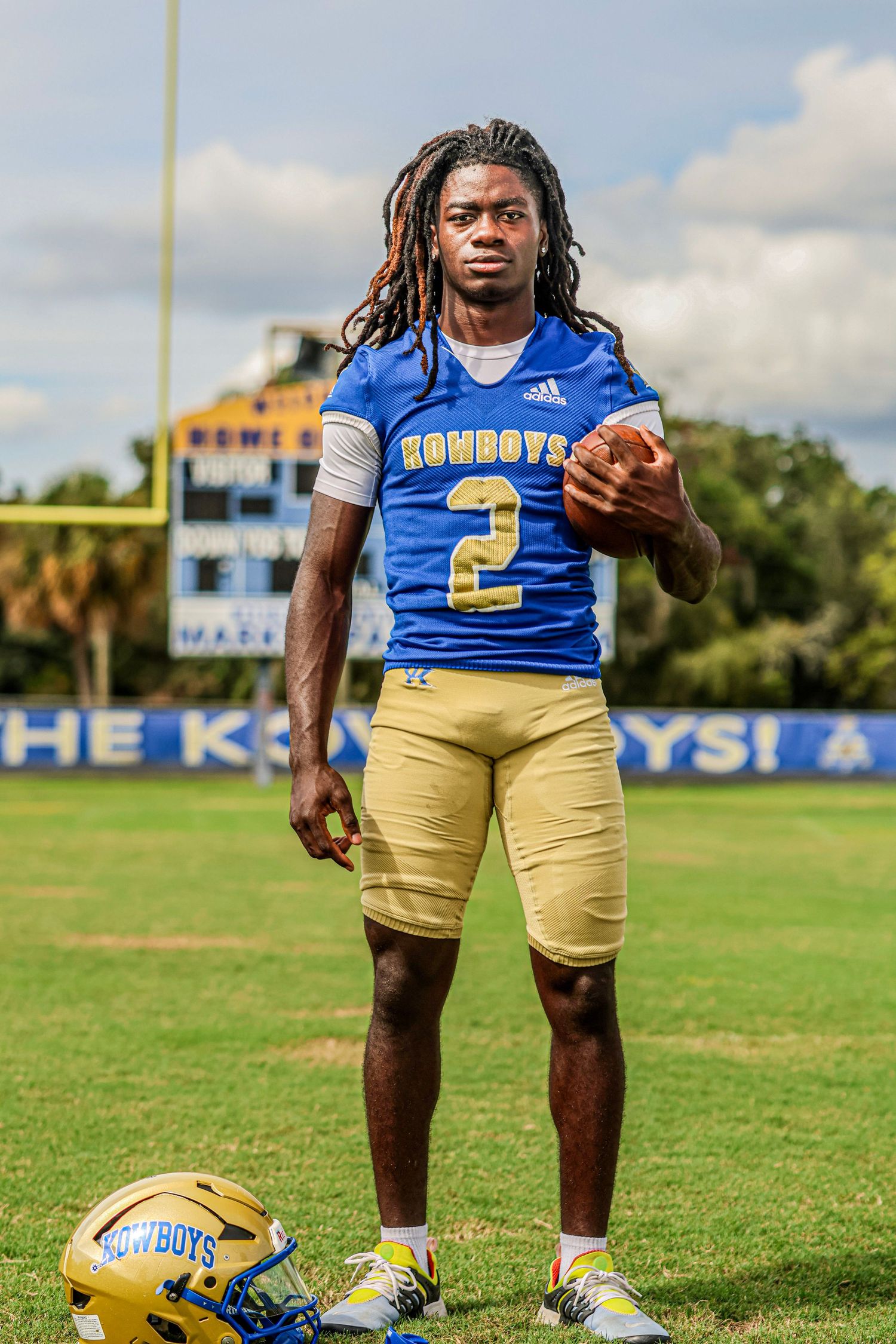 Football player in blue and gold Cowboys uniform poses on practice field with equipment and ball.