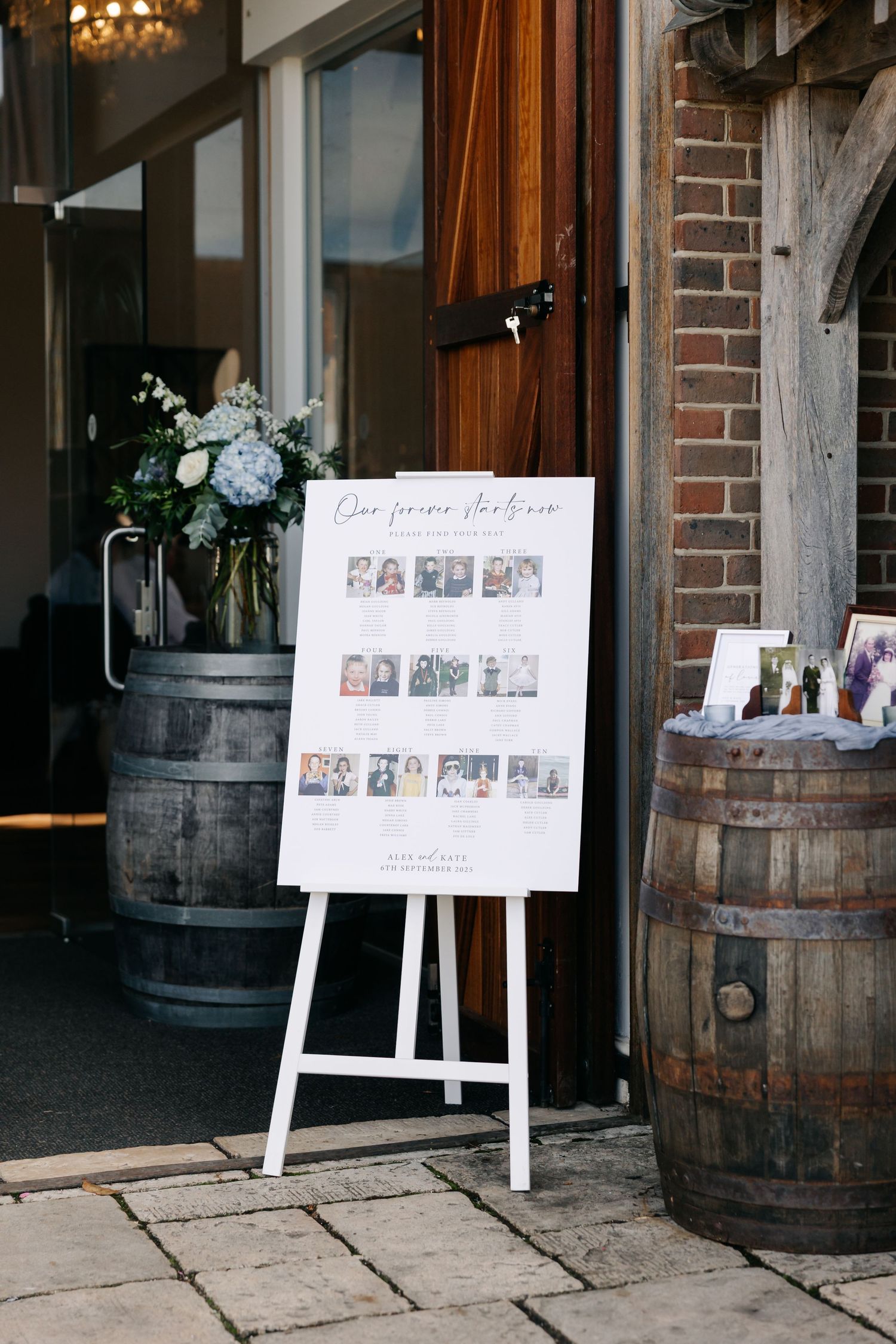 Photo displays of wedding pictures on white easels arranged near rustic wooden barrels in an indoor venue.