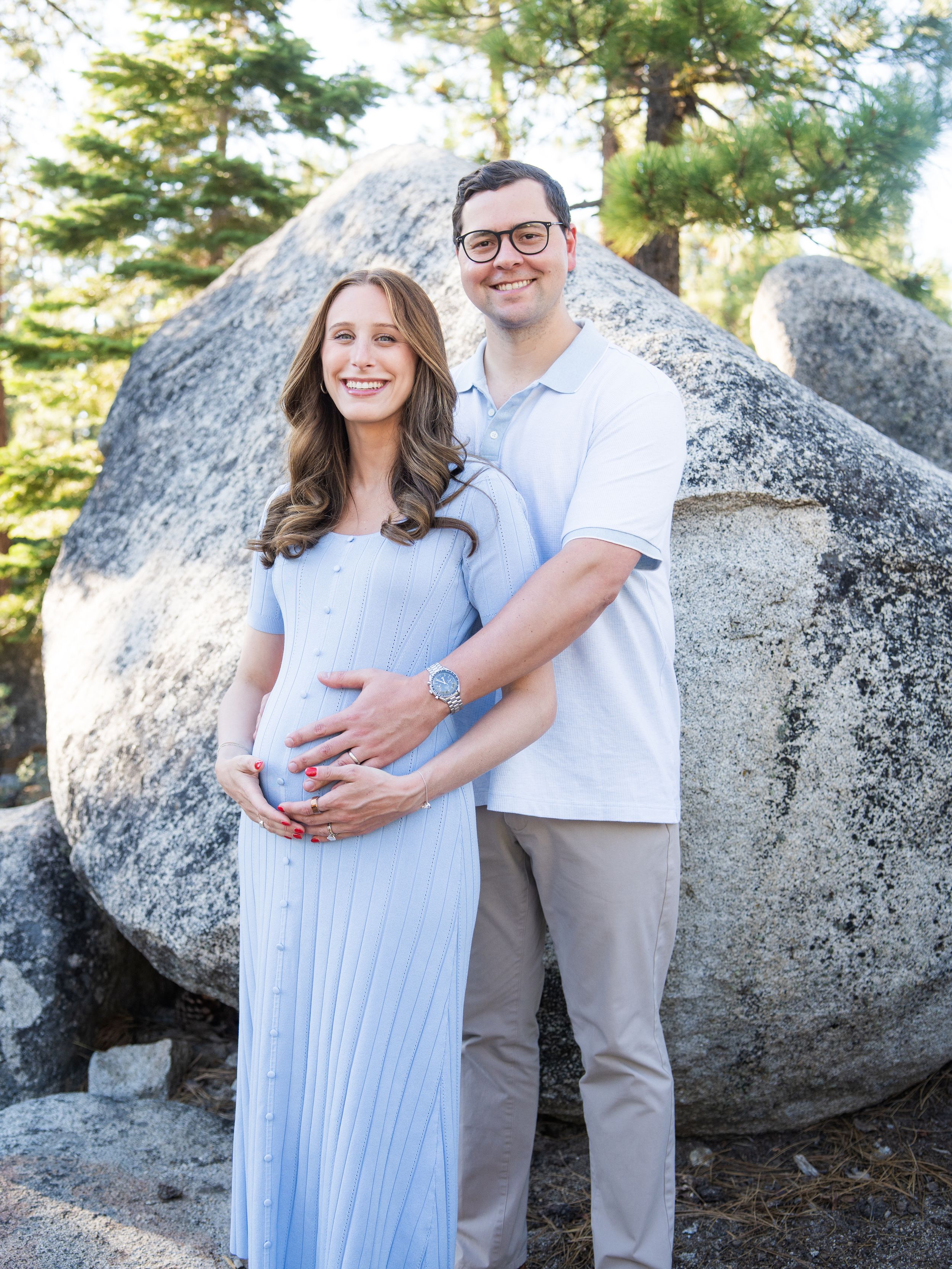 Expectant couple shares tender moments against a backdrop of large granite boulders and pine trees in outdoor maternity photos.