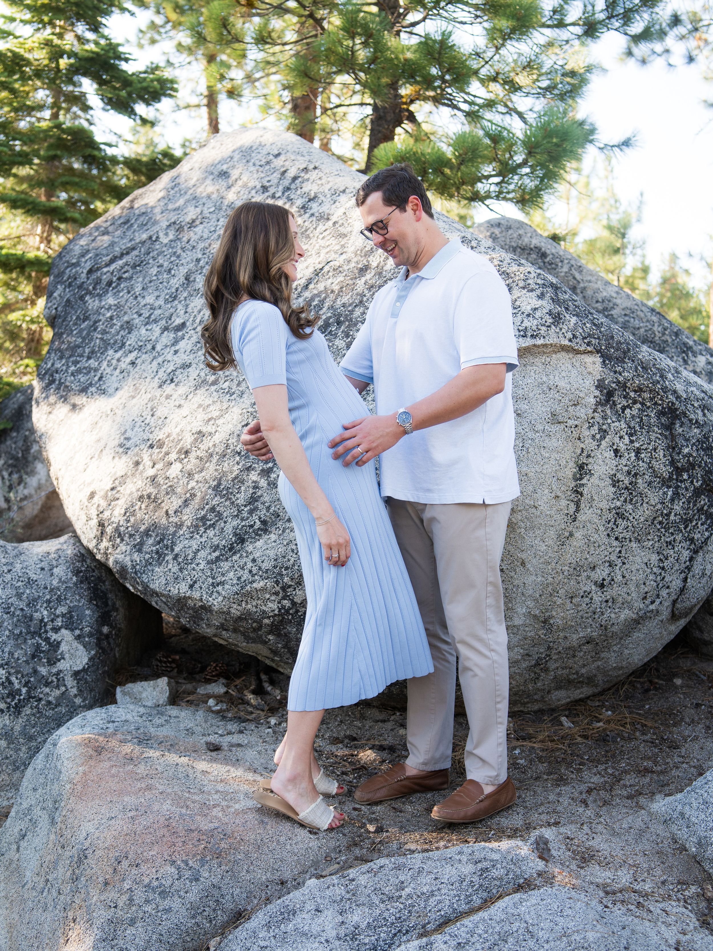 A couple stands together on large granite boulders in a pine forest setting for maternity photos.