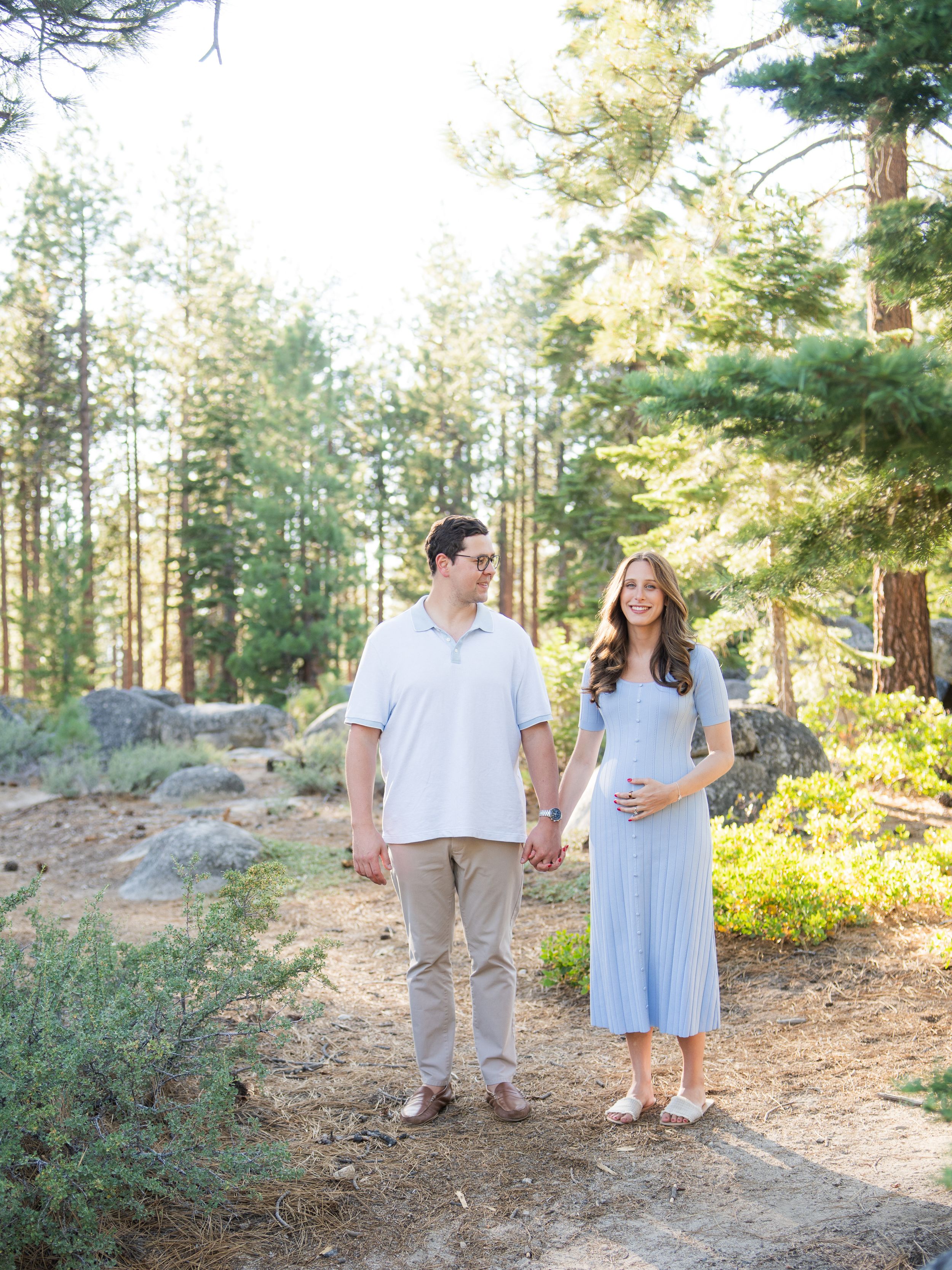 A couple walks hand in hand through a sunlit forest path among tall pine trees in matching light colored outfits.