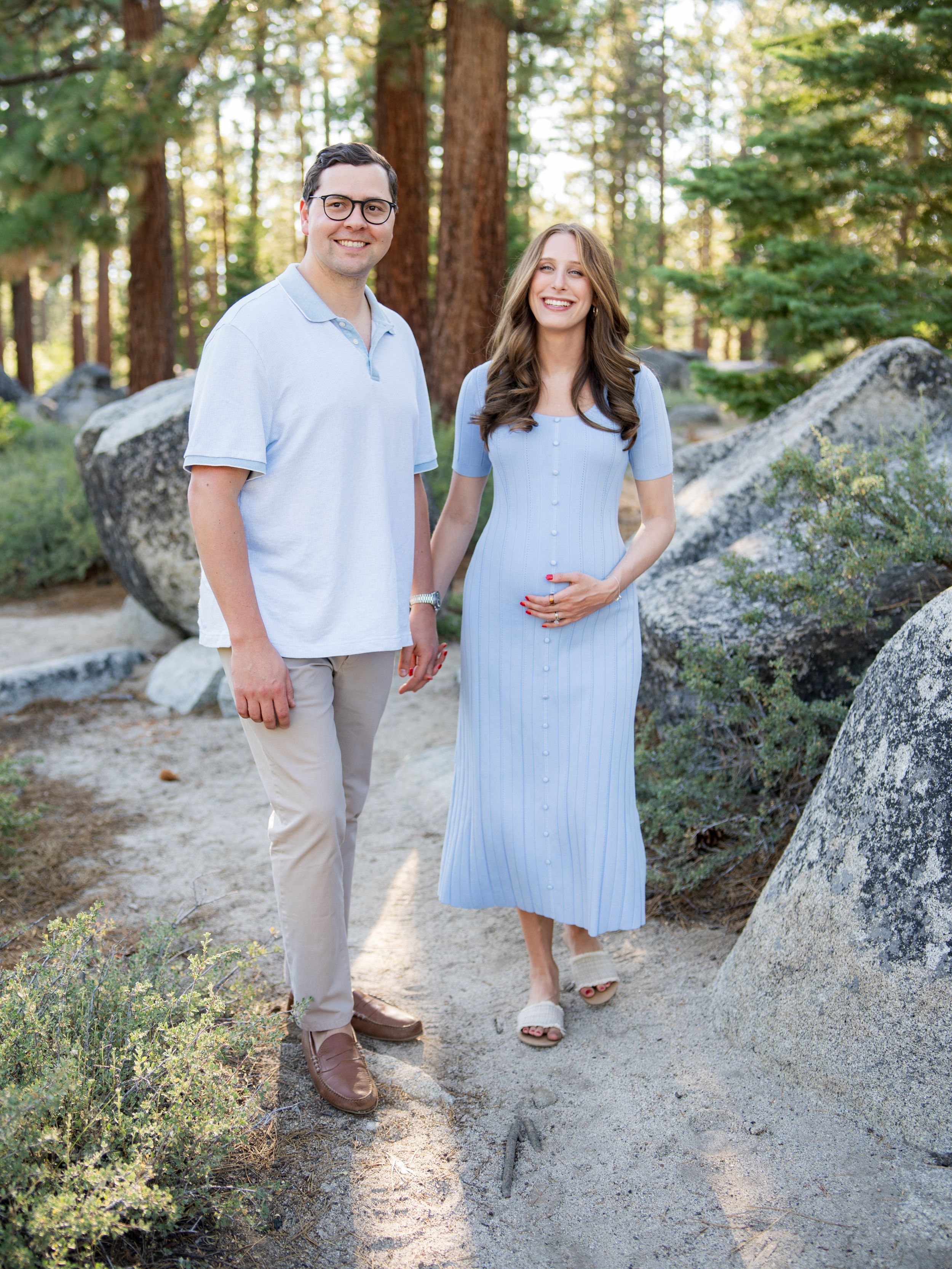 A couple stands together on a rocky forest path surrounded by tall pine trees on a sunny afternoon.