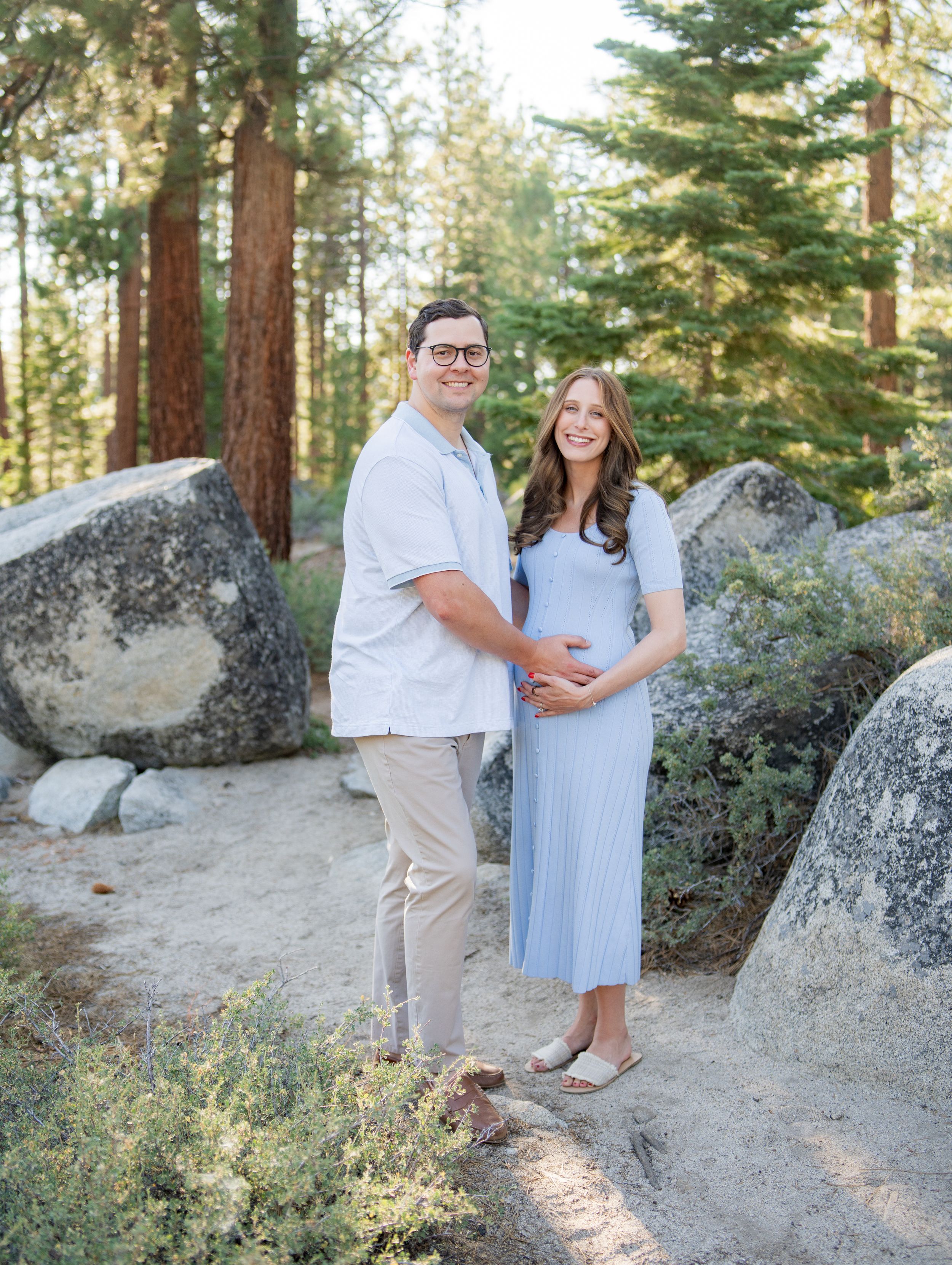 A couple poses together among tall pine trees and granite boulders in a romantic mountain forest setting.