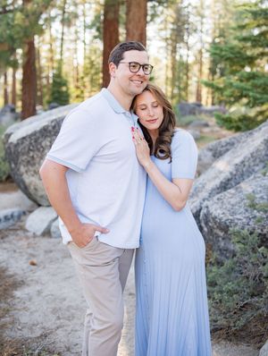 A couple shares a tender moment among large granite boulders in a sunlit forest setting wearing light blue and white.