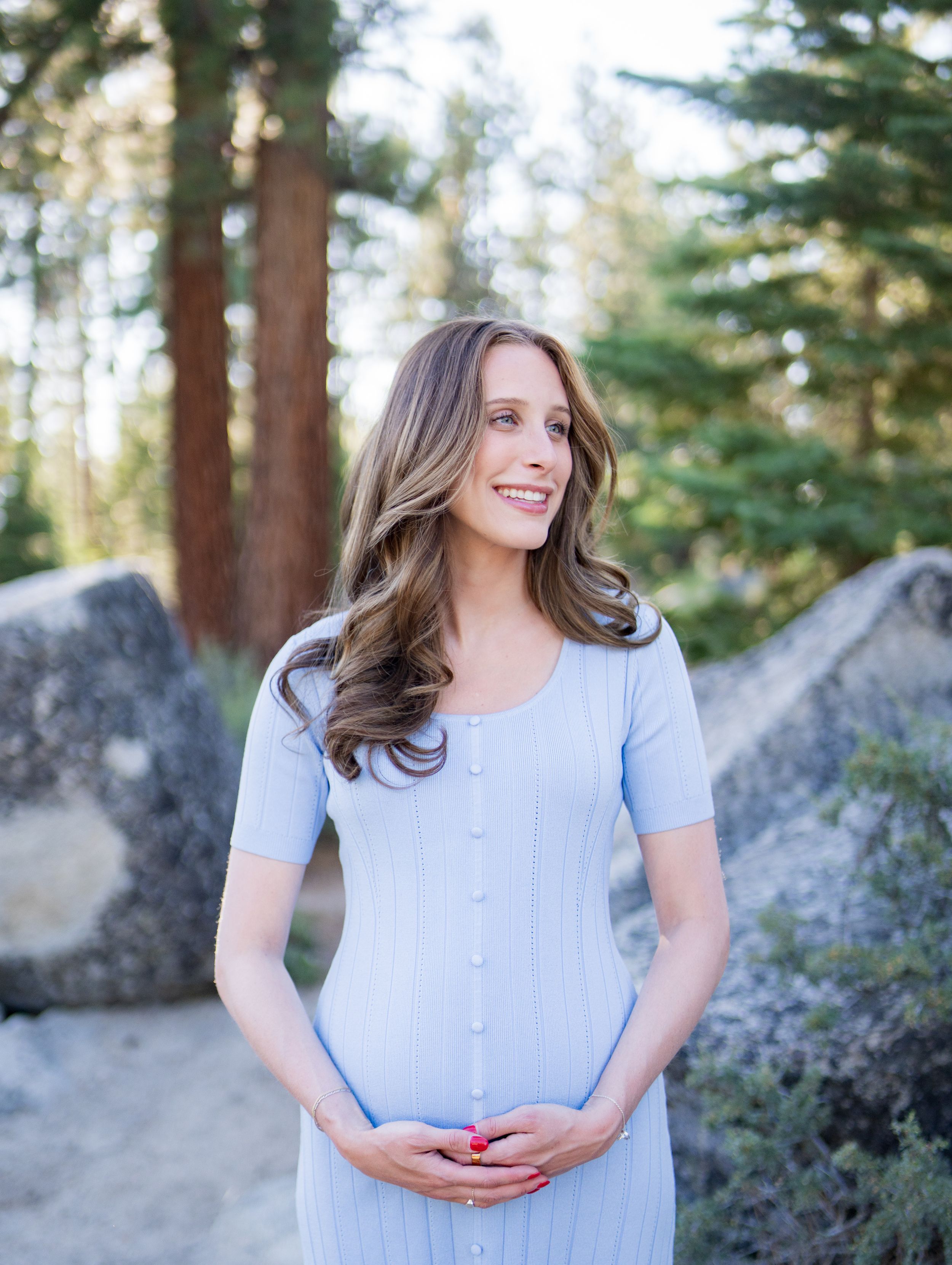 A woman in a light blue dress stands among evergreen trees and large boulders in natural lighting.