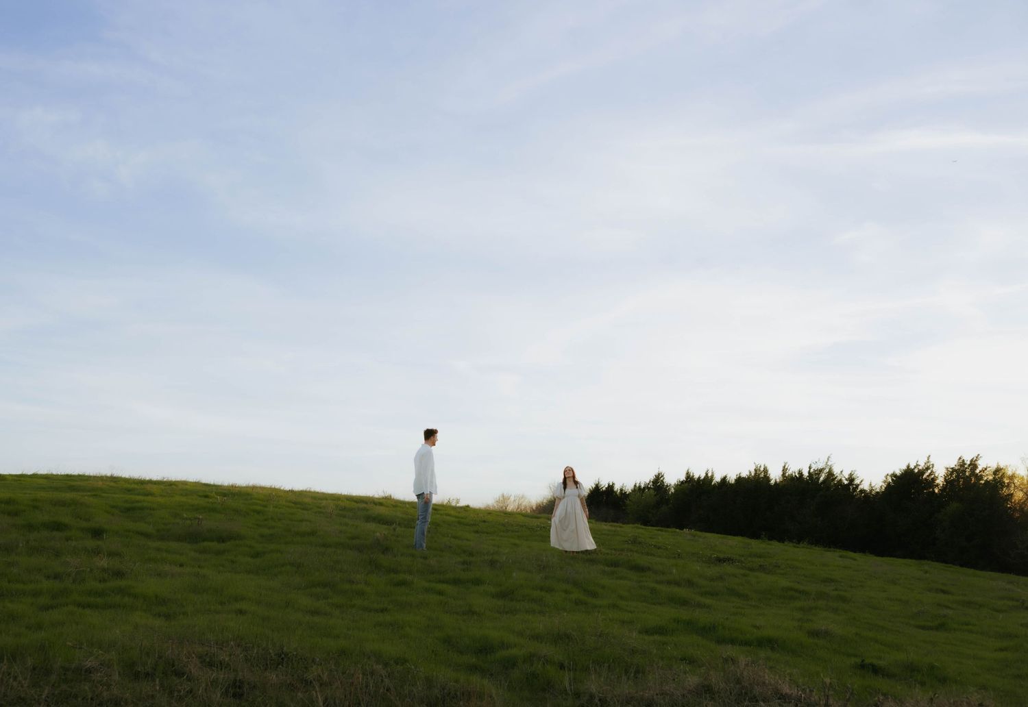 Couple standing apart on a lush green hillside under a wide open sky at golden hour.
