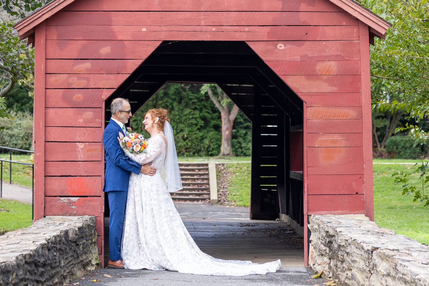 A couple shares a romantic moment under a rustic red covered bridge during their wedding photoshoot.