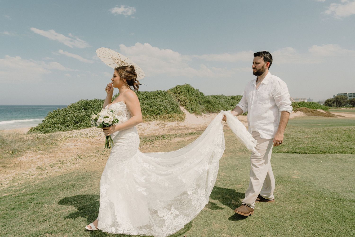 A couple in wedding attire walks together on a scenic coastal golf course overlooking the ocean on a sunny day.
