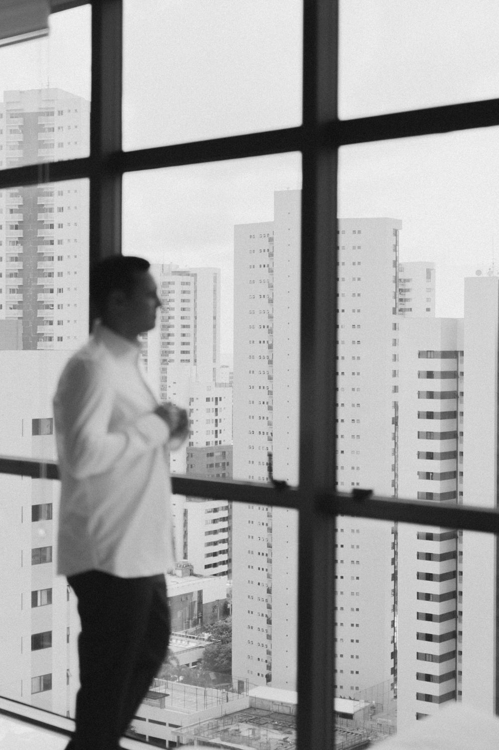 Black and white photo of a person in white shirt looking through a window at tall apartment buildings in an urban landscape.