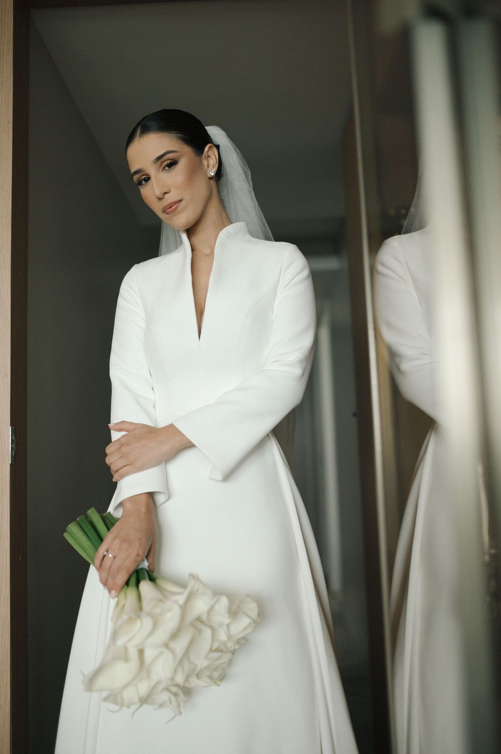 Elegant bride in a white long-sleeved gown poses in dramatic window lighting.