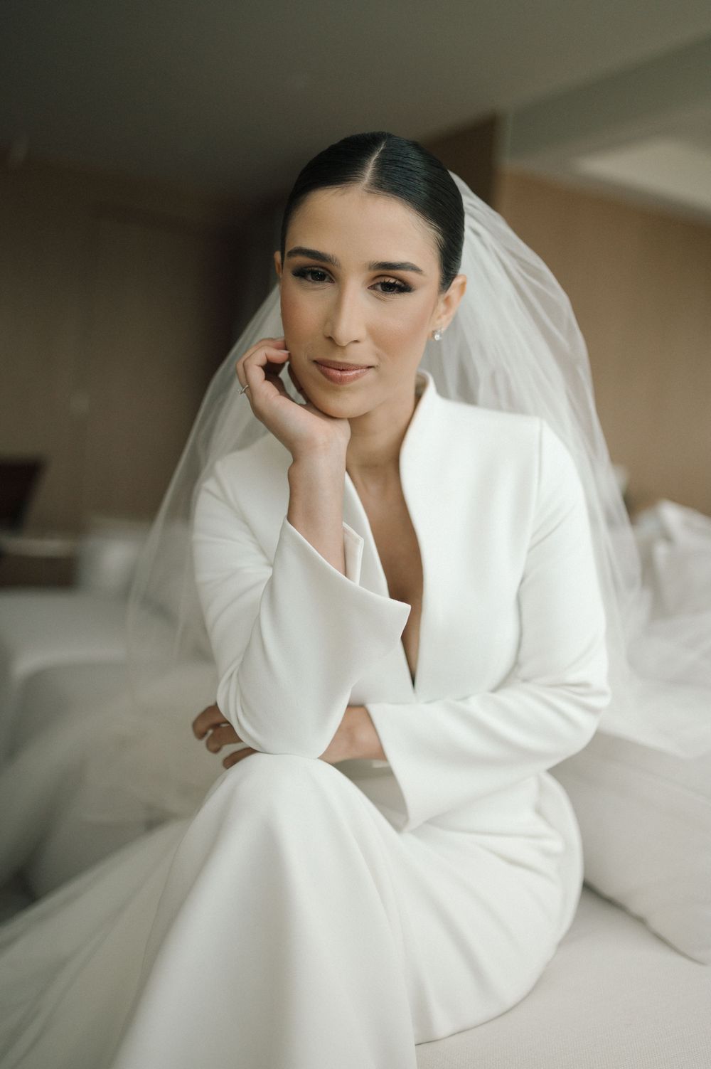 Person in white bridal attire reclining elegantly on white bedding in soft natural light.