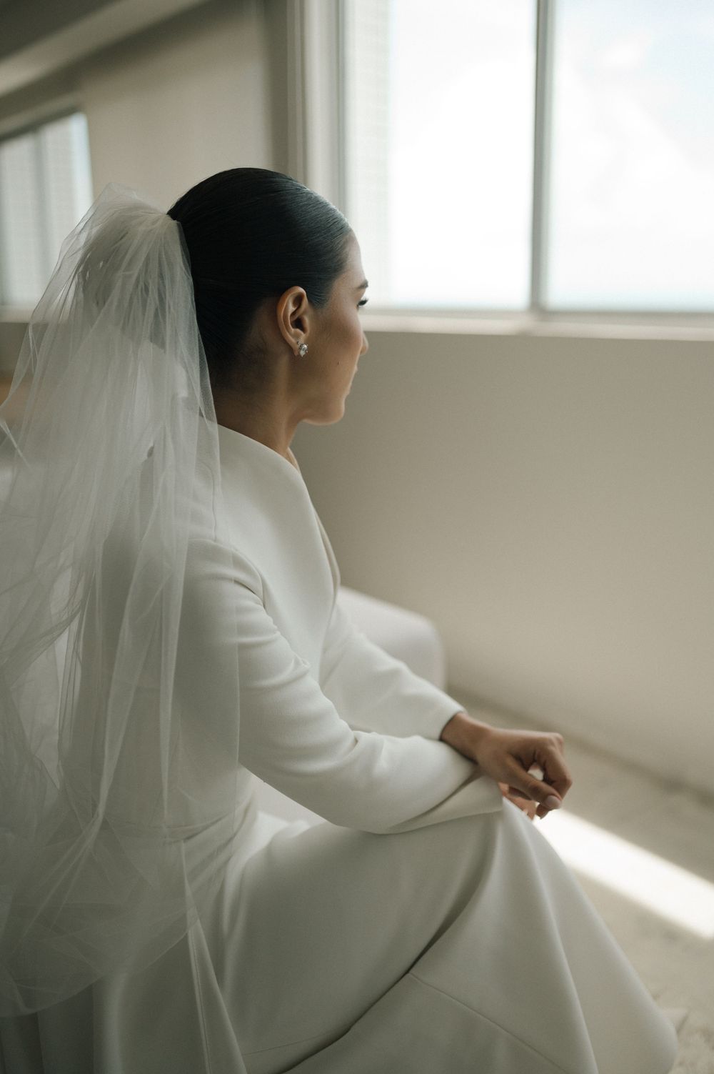 Elegant bride in a minimalist white dress sits peacefully by a window in natural light.