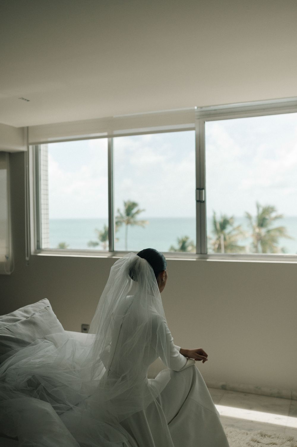 A flowing white wedding dress and veil catch the light near large windows with an ocean view.