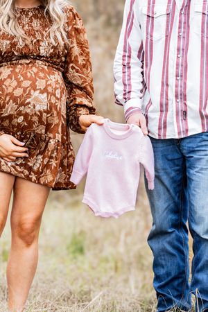 Couple holding tiny pink baby onesie while wearing brown floral dress and striped shirt in outdoor setting.