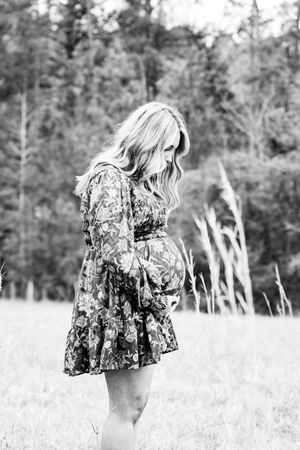Black and white photo of pregnant woman in floral dress standing in field.