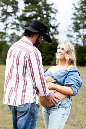 A couple in denim and stripes embrace in a sunny field with pine trees in the background.
