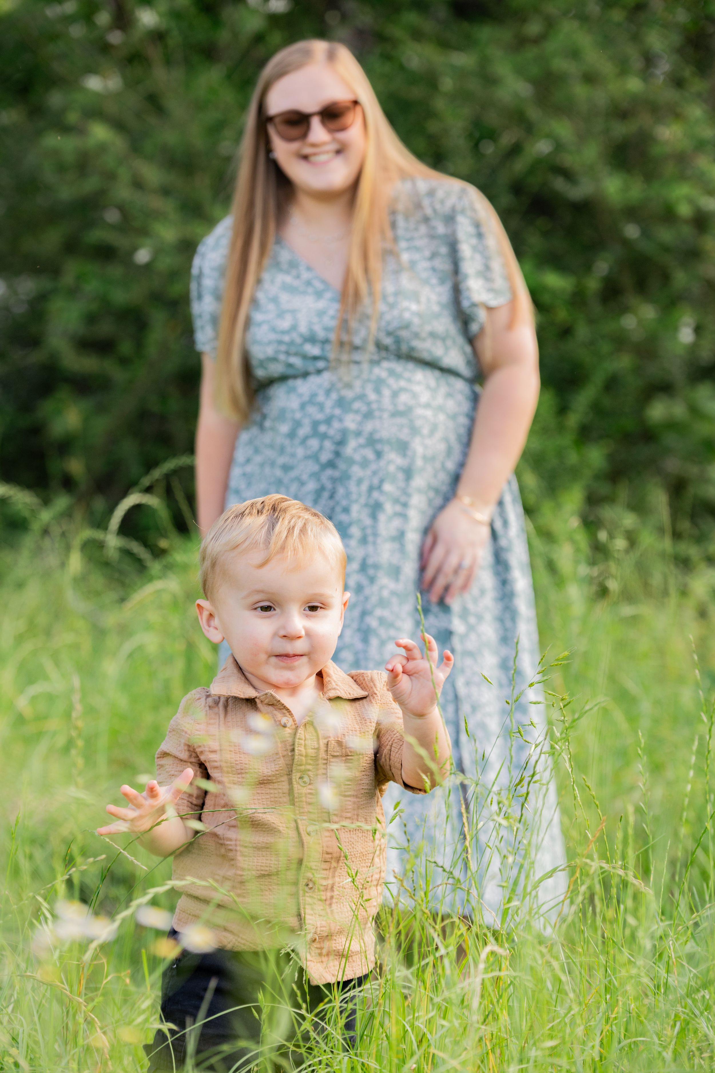 Pregnant woman in blue floral dress smiles as toddler boy plays in tall green grass outdoors.