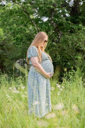 Pregnant woman in floral dress holding baby bump in sunny green meadow.