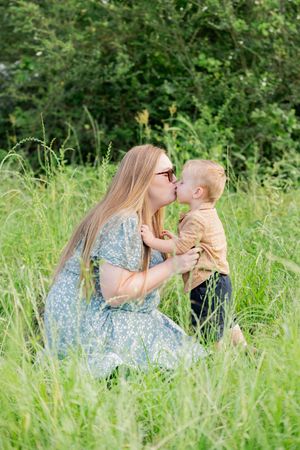 Mother in blue floral dress kisses young toddler son while sitting in lush green meadow grass.