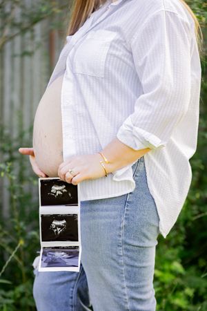 Pregnant woman in white shirt and jeans holding ultrasound photos beside her baby bump outdoors.