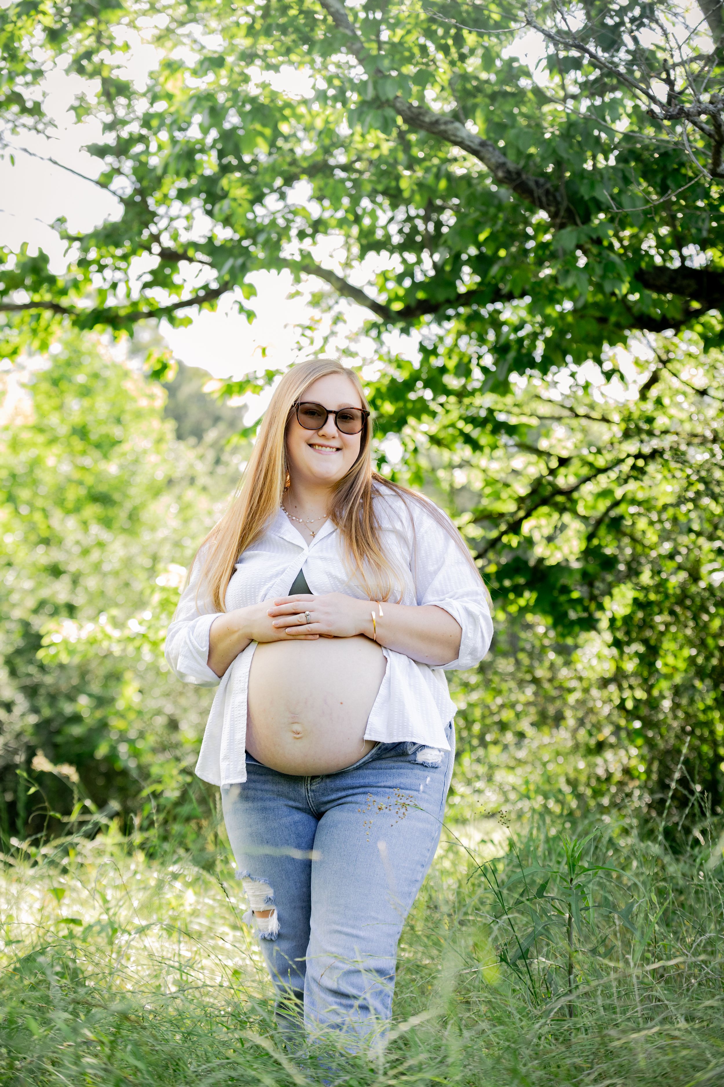 Smiling pregnant woman in white shirt and jeans cradling baby bump outdoors in lush green garden on sunny day.