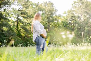 Pregnant woman in white shirt and jeans stands in sunlit meadow cradling her baby bump outdoors.