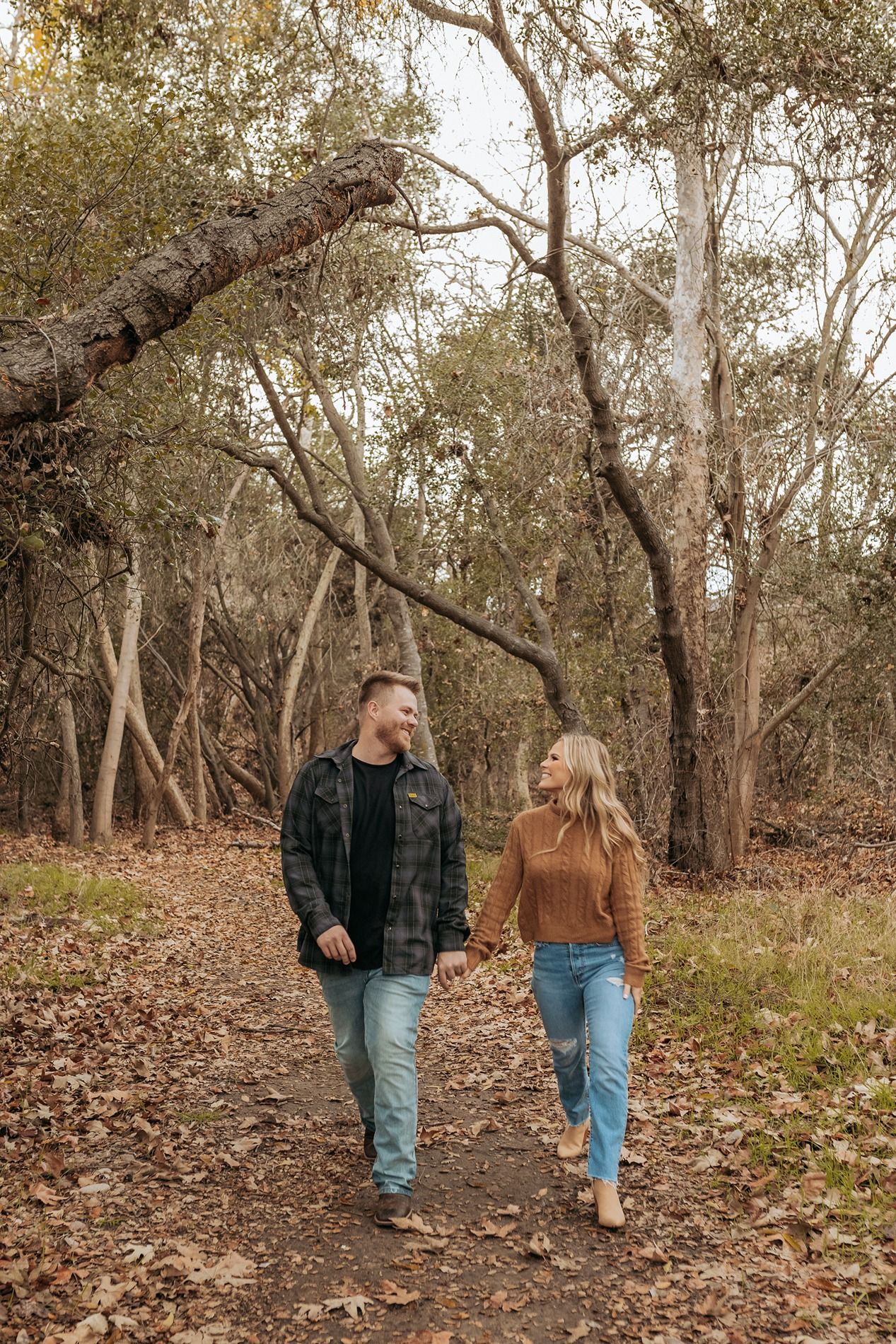 A couple walks together on a leaf-covered trail through autumn woods wearing casual attire in San Diego.