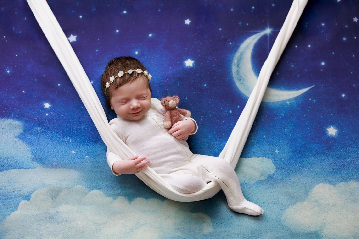 Photo of a smiling newborn baby girl sitting on a swing with a moon and stars backdrop