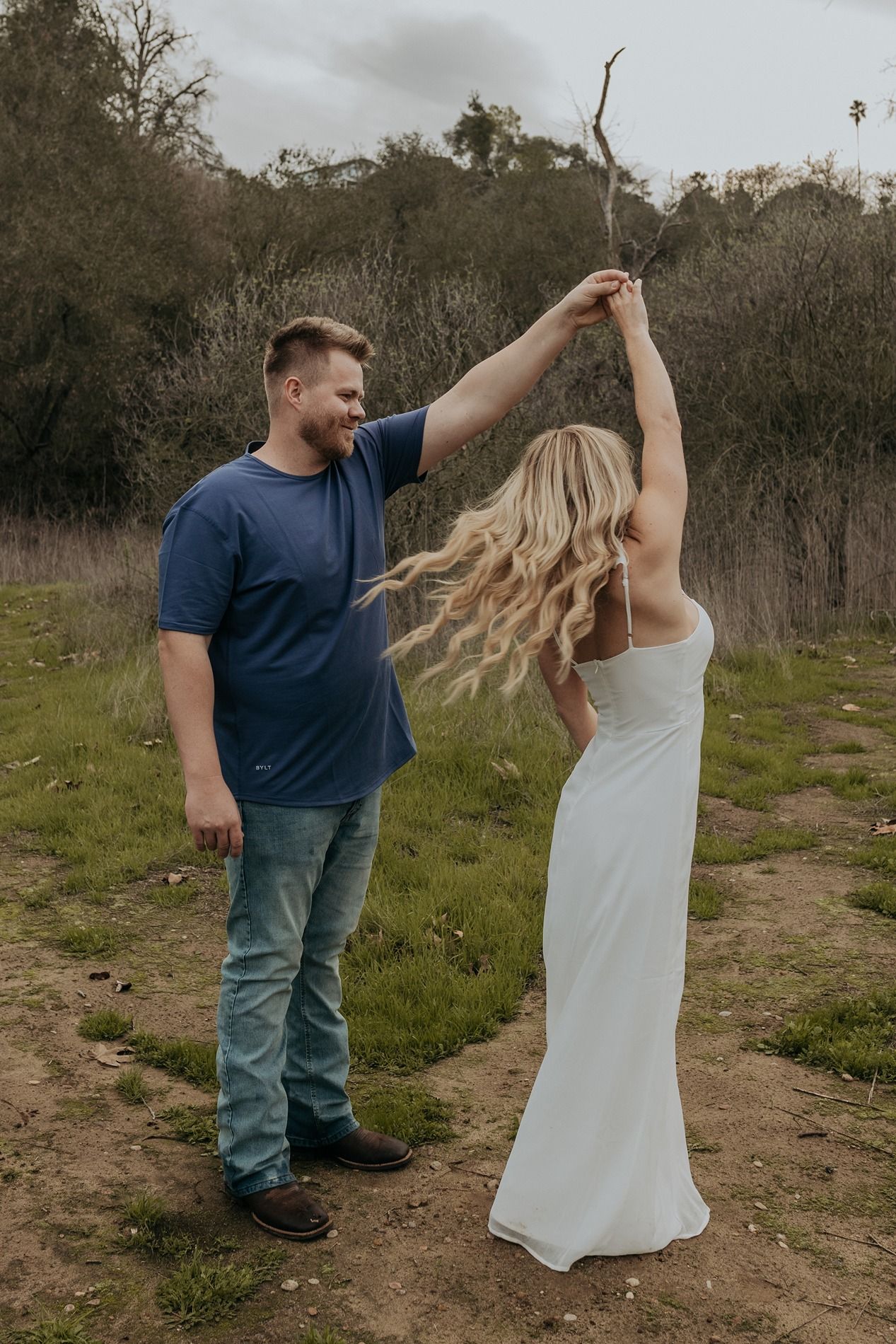 A couple in casual attire dances together on a dirt path surrounded by green vegetation and trees in San Diego.
