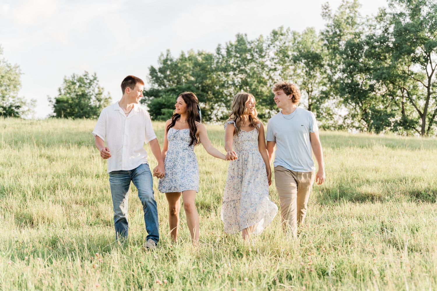 A group of high school seniors in light colored clothing walk through a sunlit meadow during a summer photo session in Toledo, Ohio