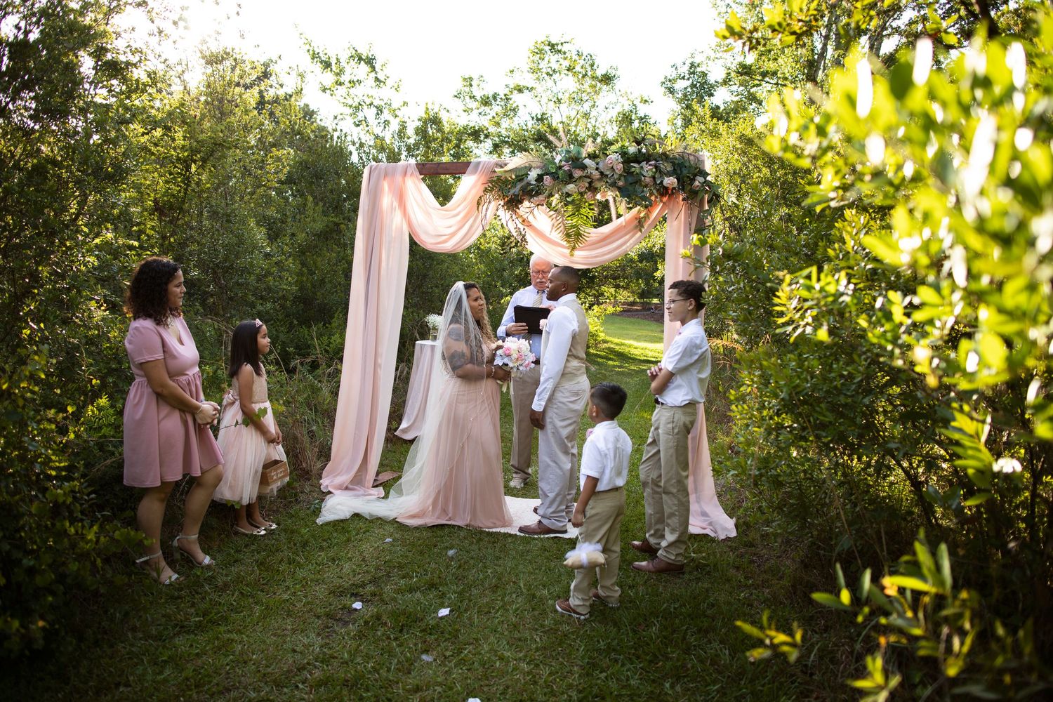Wedding party in pink dresses poses together in a lush garden setting during golden hour sunlight.
